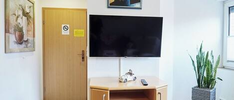 A room with a wooden cabinet, a flat-screen TV mounted on the wall, a door with a no-smoking sign, and a potted plant.
