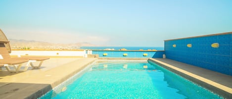 A swimming pool with clear blue water, surrounded by a blue tiled wall and a white building with a flat roof.