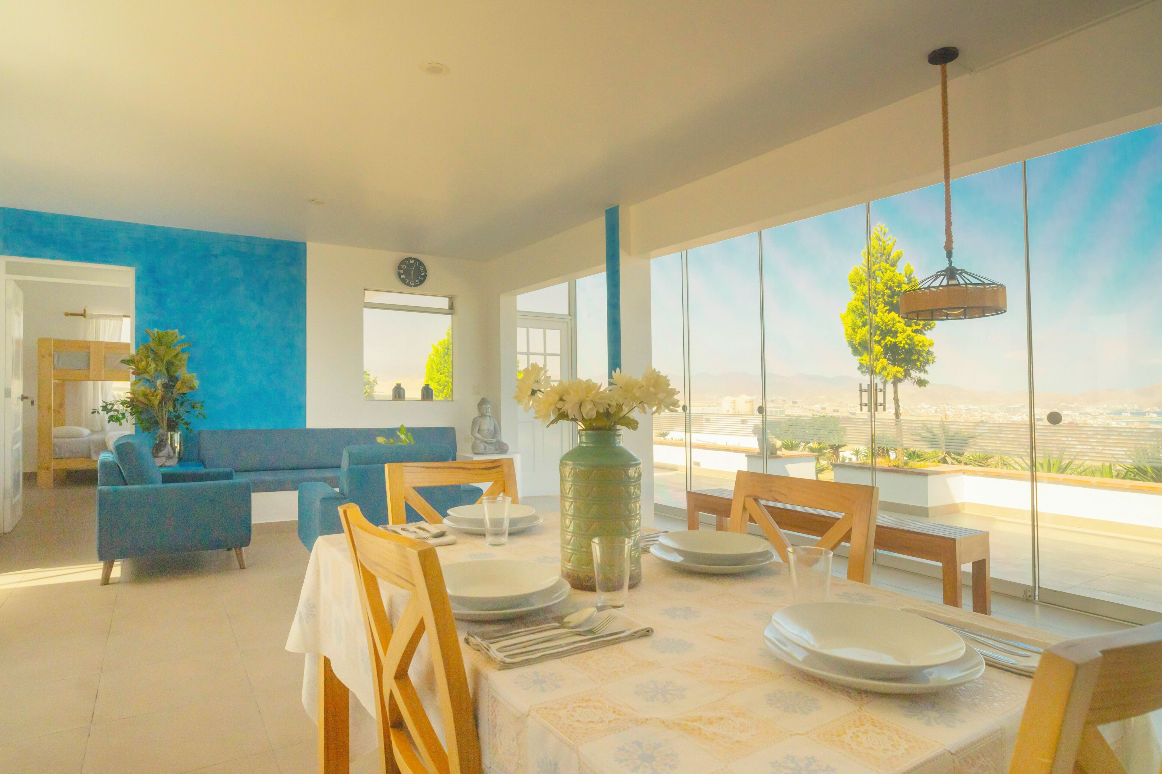 Un espace de repas moderne avec une table dressée pour deux personnes, un canapé bleu et une vue sur le paysage urbain à travers de grandes fenêtres.