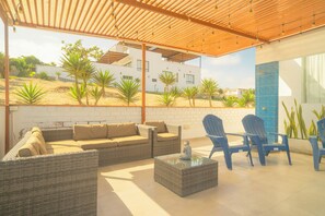 A covered outdoor seating area with wicker furniture, a glass coffee table, and a view of a modern building and palm trees.