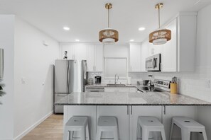 A modern kitchen with white cabinetry, a central island with bar stools, stainless steel appliances, and pendant lighting. - Brightwater Beach Condo | Bay Views & Beach Access (St. Petersburg)