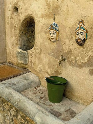 Eine Wand mit einem kleinen Rundbogenfenster, zwei Schmuckmasken, einem Messinghahn und einem grünen Eimer.