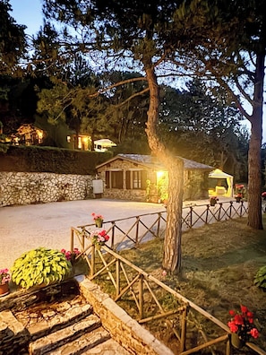 A stone-walled building with a wooden fence and a small porch, surrounded by trees and a well-lit garden area. - Stone cottage with view of Gubbio and shared pool (Gubbio)
