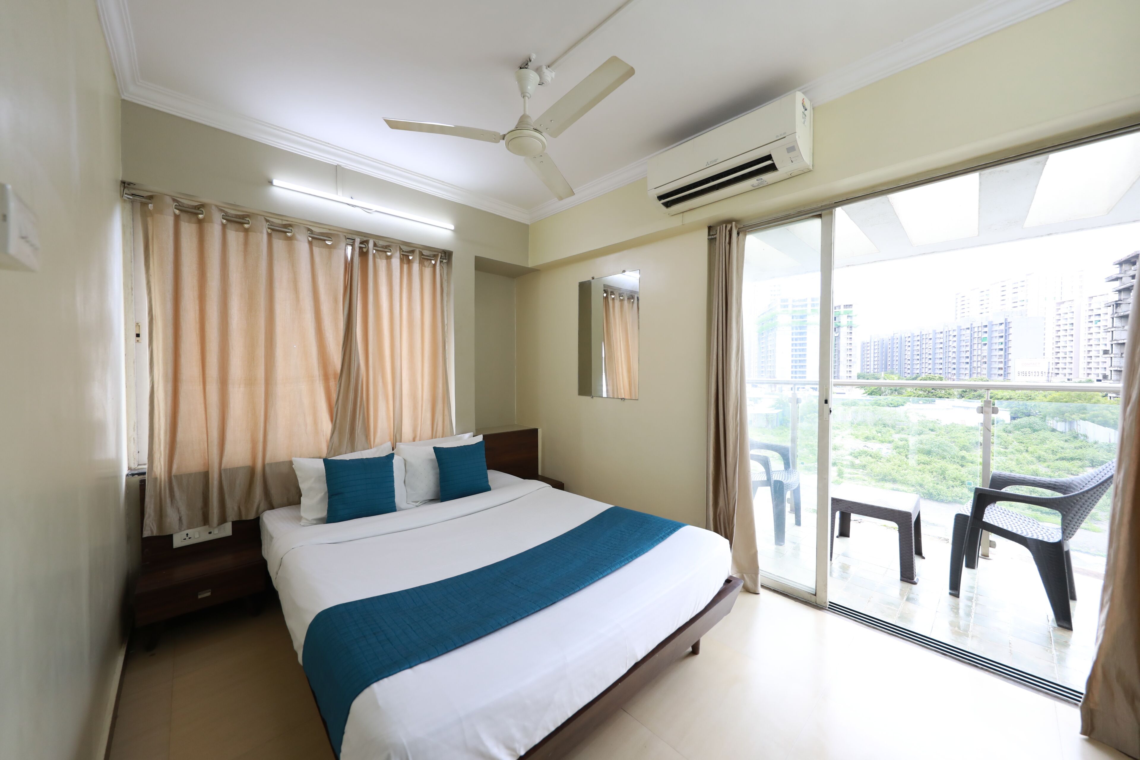 A hotel room with a bed, a ceiling fan, an air conditioning unit, a balcony with chairs, and a view of buildings.