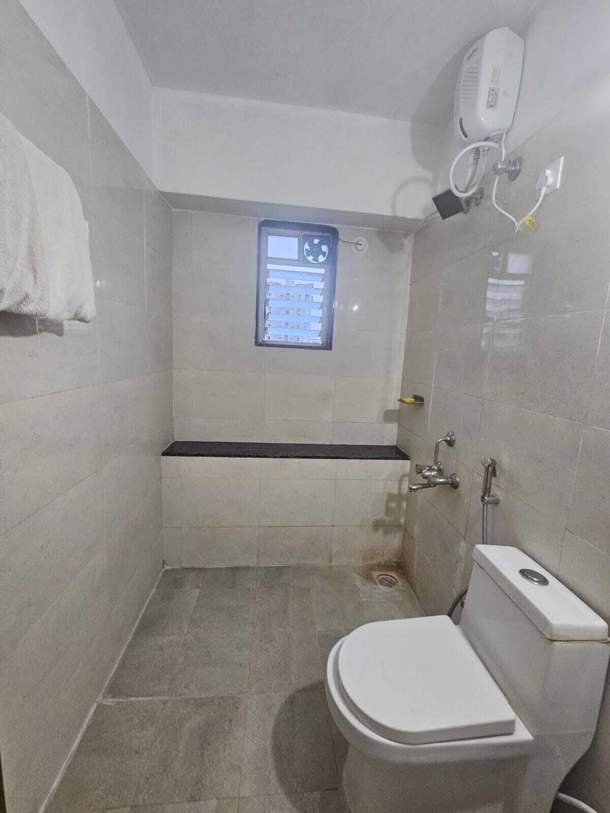 A small, tiled bathroom with a toilet, a shower, and a window with a control panel.