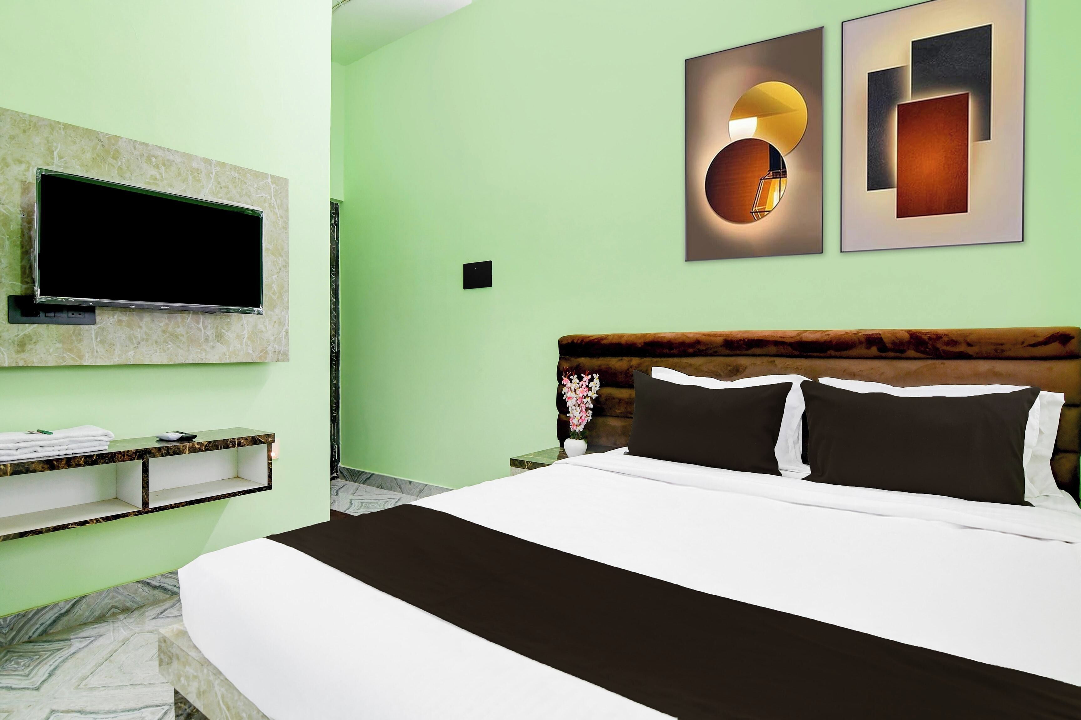 A hotel room with a bed, a flat-screen TV mounted on the wall, a small shelf with towels, and a wall-mounted light fixture.