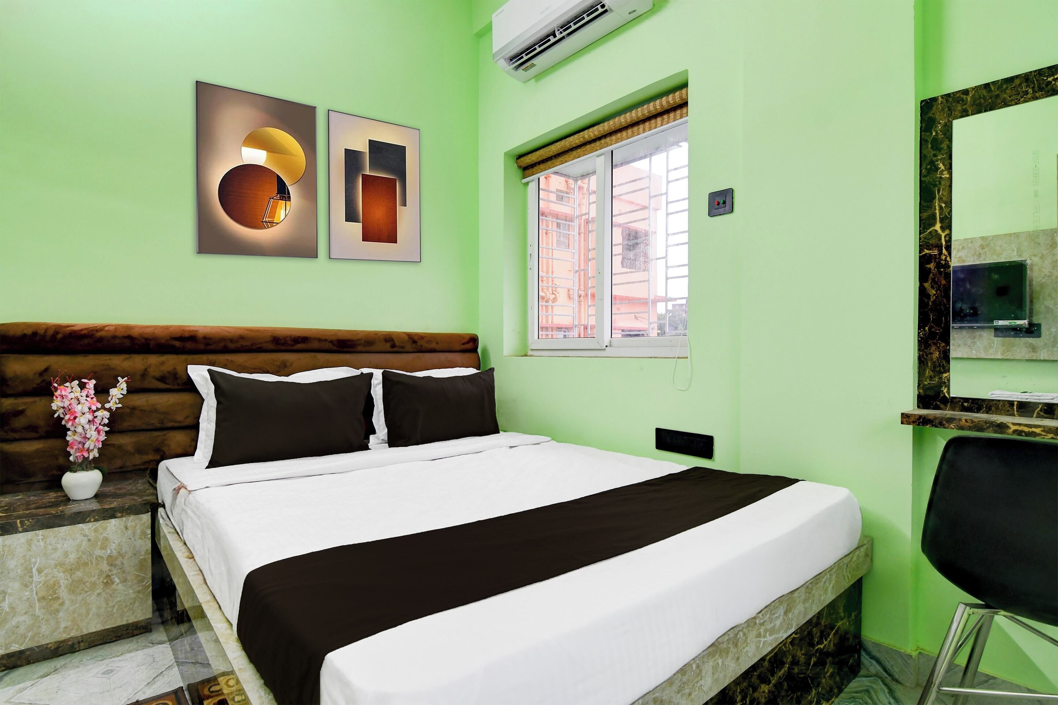 A hotel room with a bed, a nightstand, a vase with flowers, a window with a view, and a wall-mounted air conditioner.