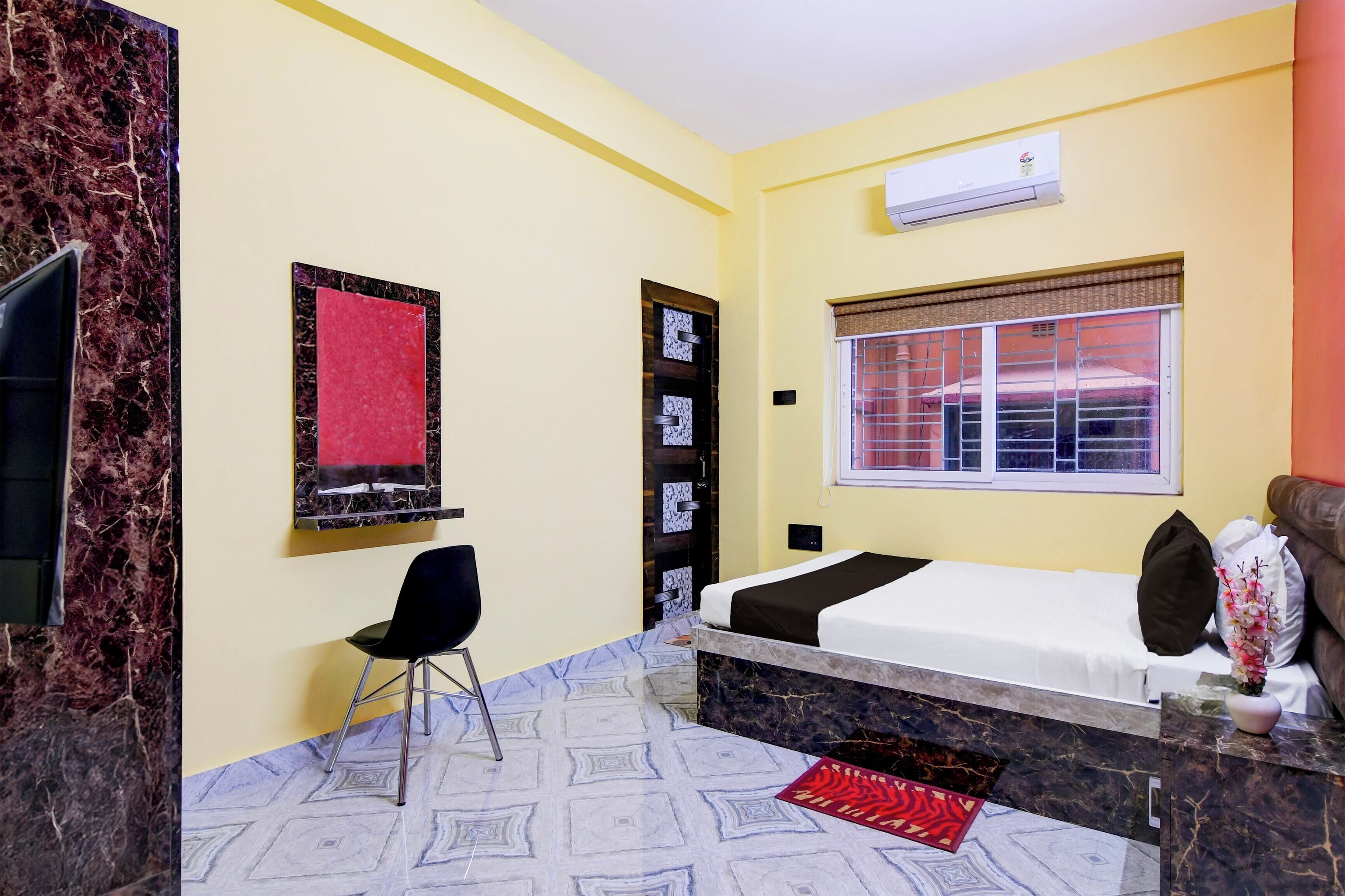 A hotel room with a bed, a chair, a window with blinds, a wall-mounted air conditioner, and a decorative vase.