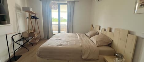 A bedroom with a wooden bed, a small bedside table with a lamp, a window with curtains, and an air conditioning unit on the wall.