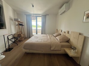 Et soveværelse med en træseng, et lille sengebord med lampe, et vindue med gardiner og en aircondition-enhed på væggen.