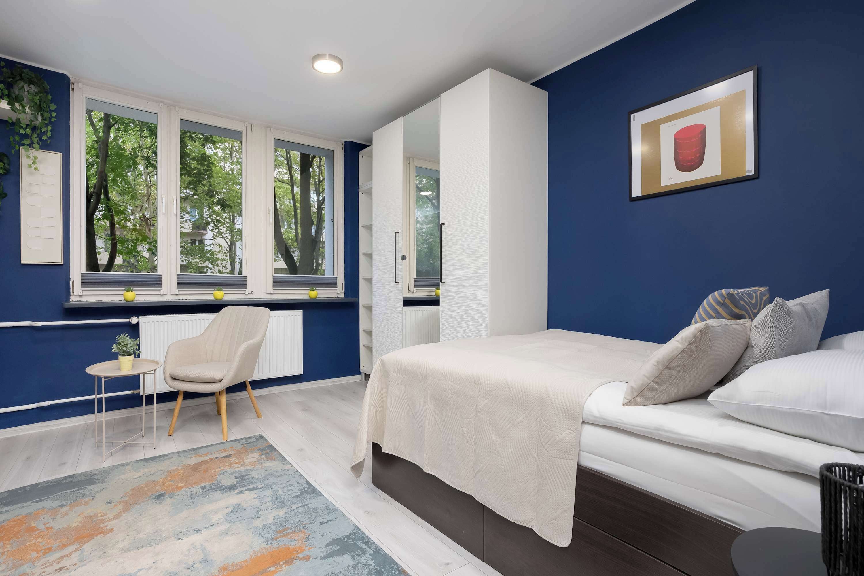 Un dormitorio con cama, una silla, una mesita y una ventana que da a árboles.