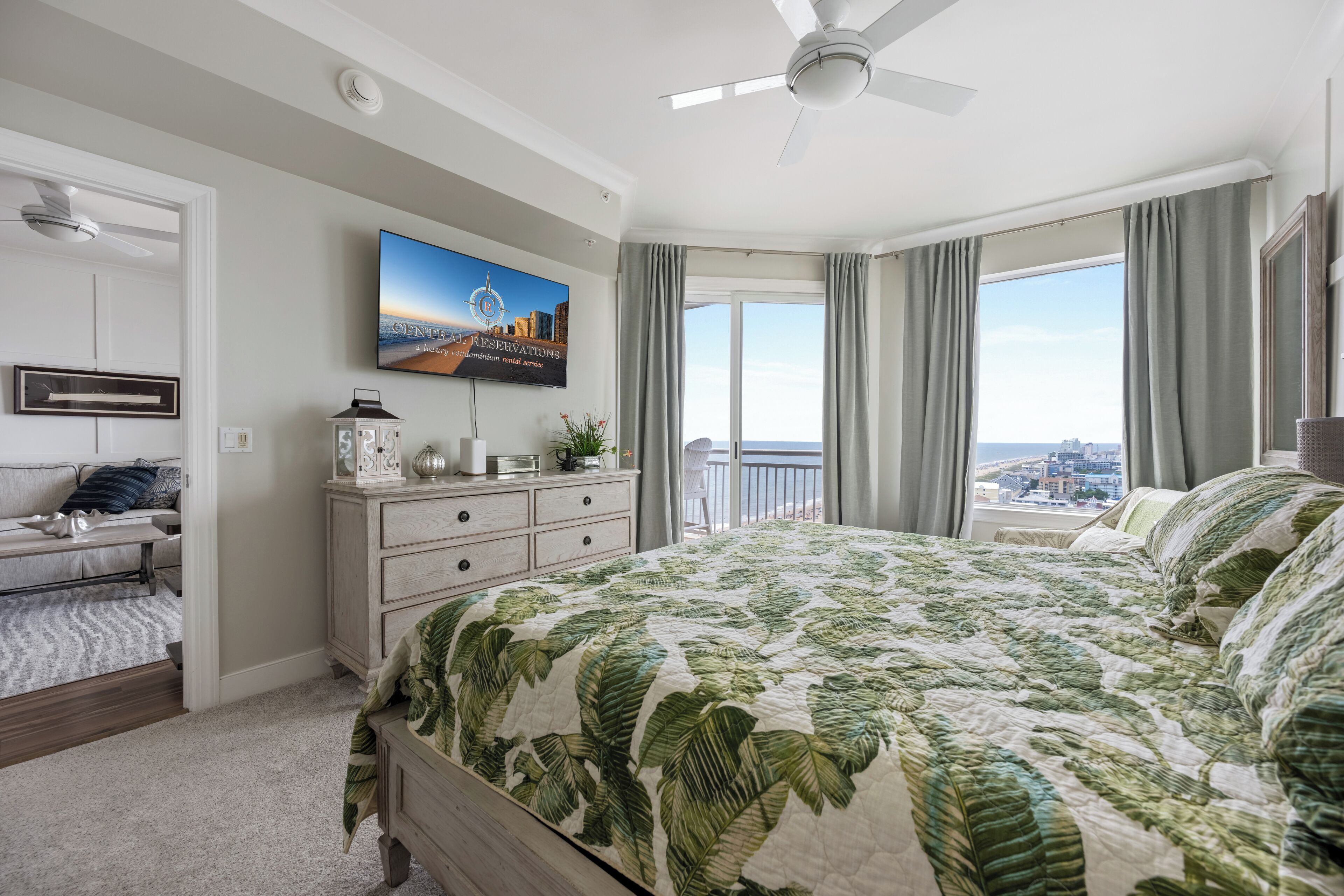 Ein Schlafzimmer mit einem großen Bett, einem Deckenventilator, Blick auf die Stadt durch das Fenster und einem an der Wand befestigten Fernseher.