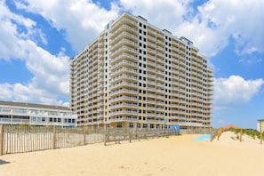 Un hôtel à plusieurs étages, doté de balcons, situé près d’une plage, avec une clôture en bois et un sentier sablonneux.