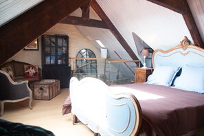Ein Schlafzimmer mit schrägem Dach, einem Bett, einem Nachttisch, einer Kommode, einem Sessel und einem Bücherregal.