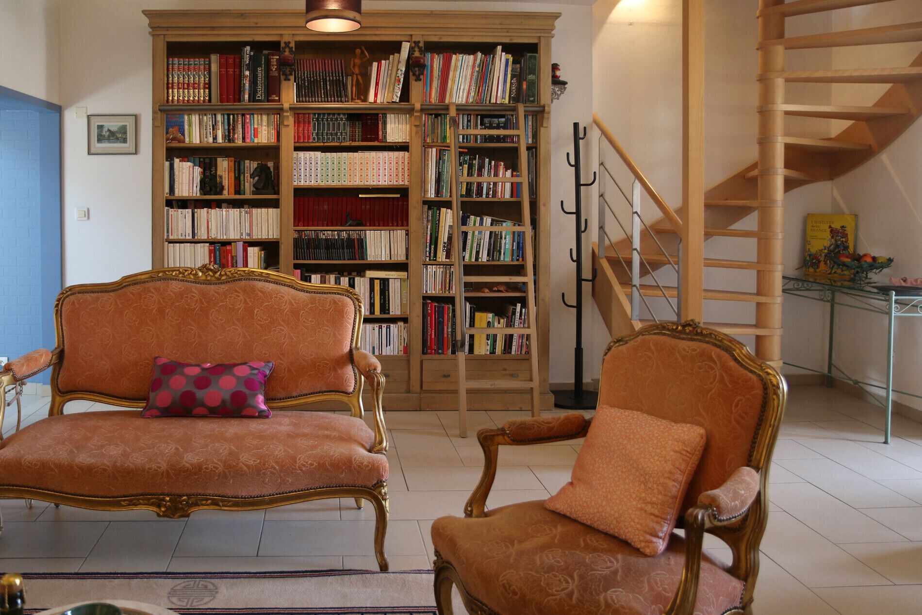 Une pièce comprenant deux fauteuils richement décorés, une grande bibliothèque et un escalier en bois.
