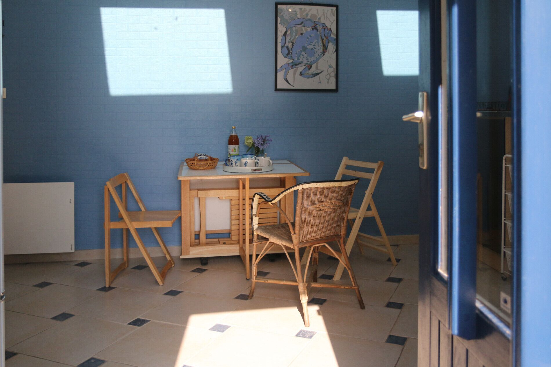 Una habitación con pared azul, una mesa de madera con sillas, un cuadro enmarco y una ventana que deja entrar luz.