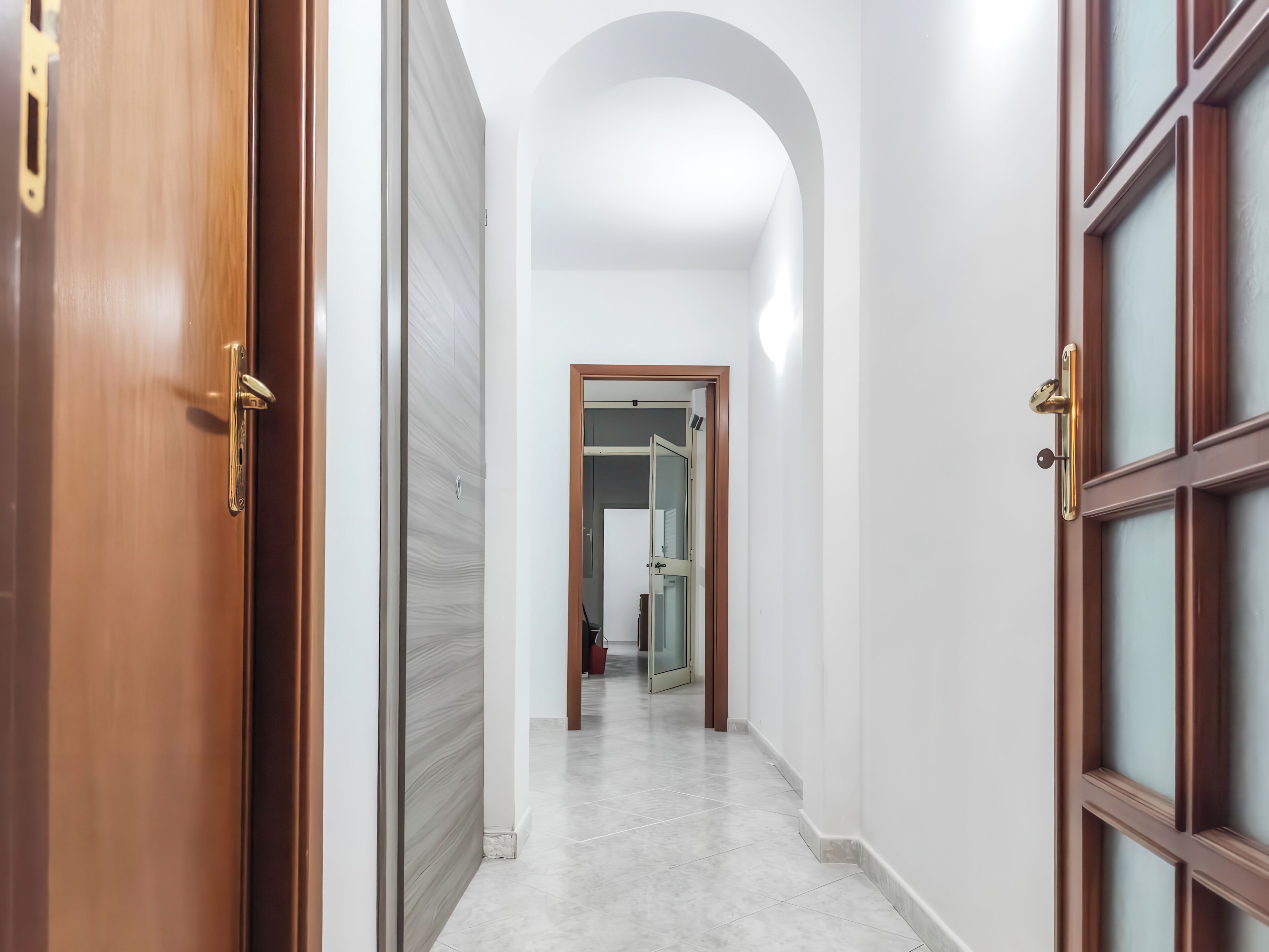 A corridor with a wooden door on the left, a white wall on the right, and a glass door at the end.
