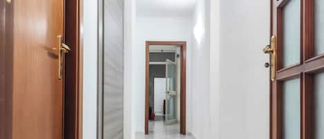 Un pasillo con una puerta de madera a la izquierda, una pared blanca a la derecha y una puerta de cristal al final.