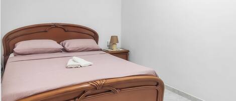 A neatly made bed with a headboard, a bedside table with a lamp, and a wall-mounted light switch.
