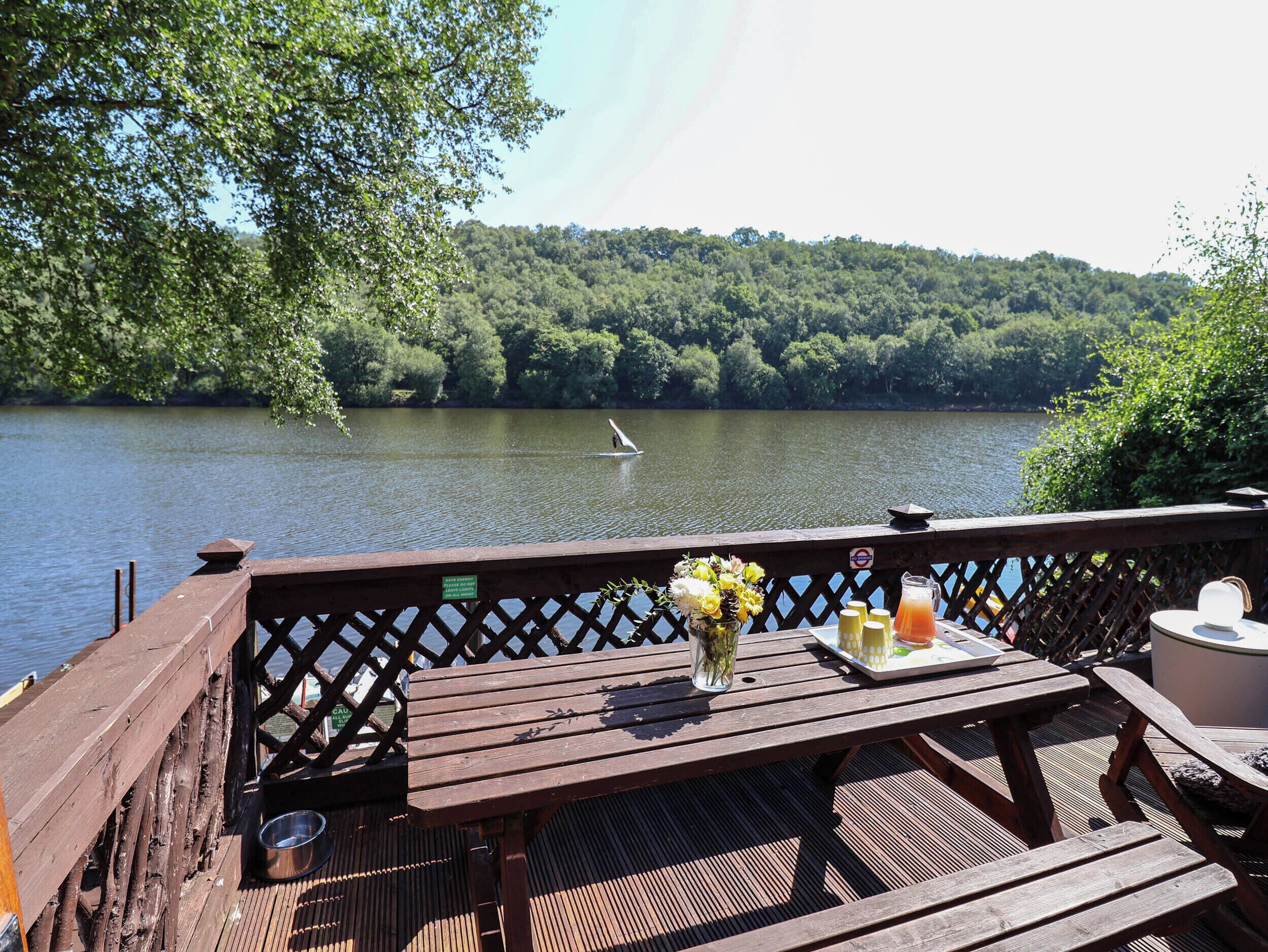 A wooden deck with a table set for dining, overlooking a calm lake with a paddleboarder in the distance.