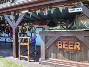 Ein rustikaler Außenbereich mit Bar, ein Holzschild mit der Aufschrift 'BEER' und ein Schild mit 'HAPPY HOUR' darüber.