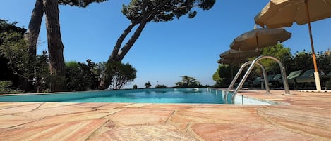 A swimming pool with stone tiles, surrounded by trees and a clear sky.