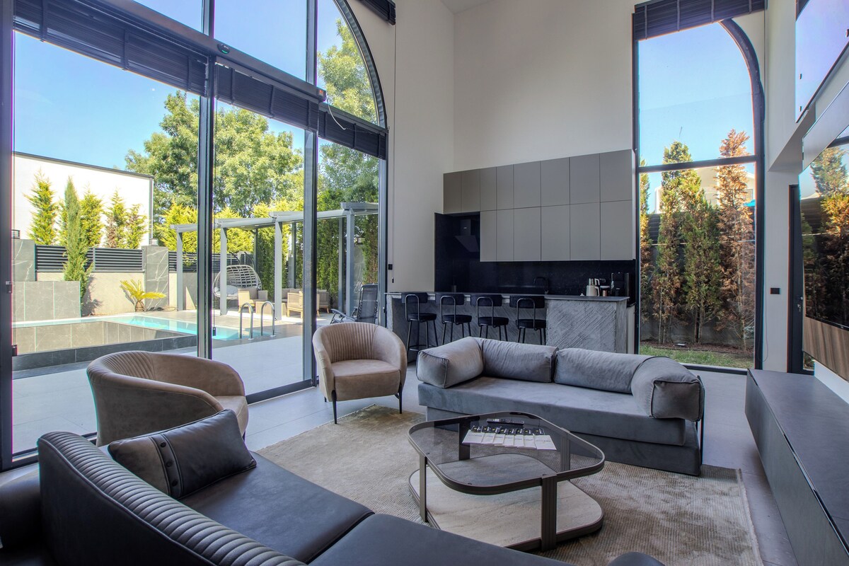A modern living room with a sofa, armchairs, and a coffee table. There is a kitchen area with cabinets and a dining set. Large windows provide a view of the outdoors.