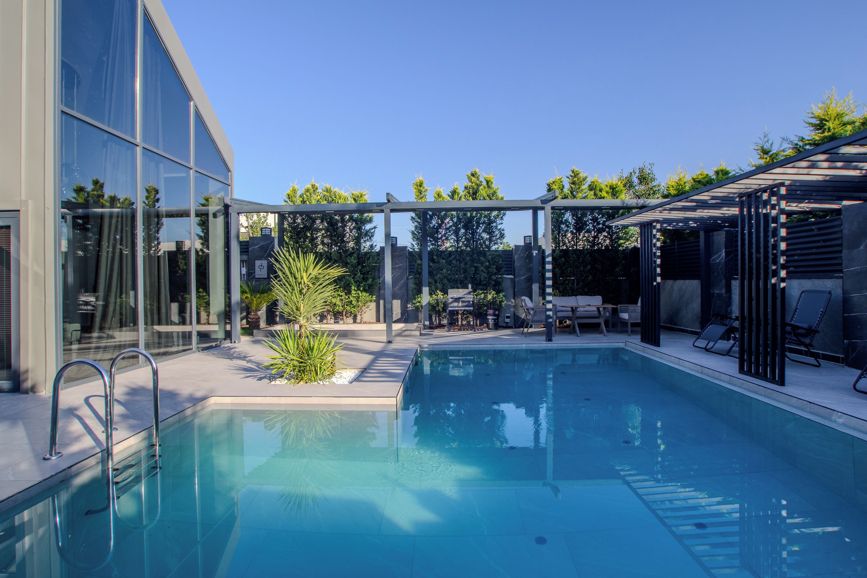 A modern outdoor pool area with a glass enclosure, lounge chairs, and a barbecue setup.
