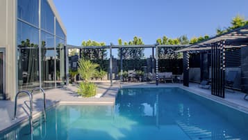 A modern outdoor pool area with a glass enclosure, lounge chairs, and a barbecue setup.