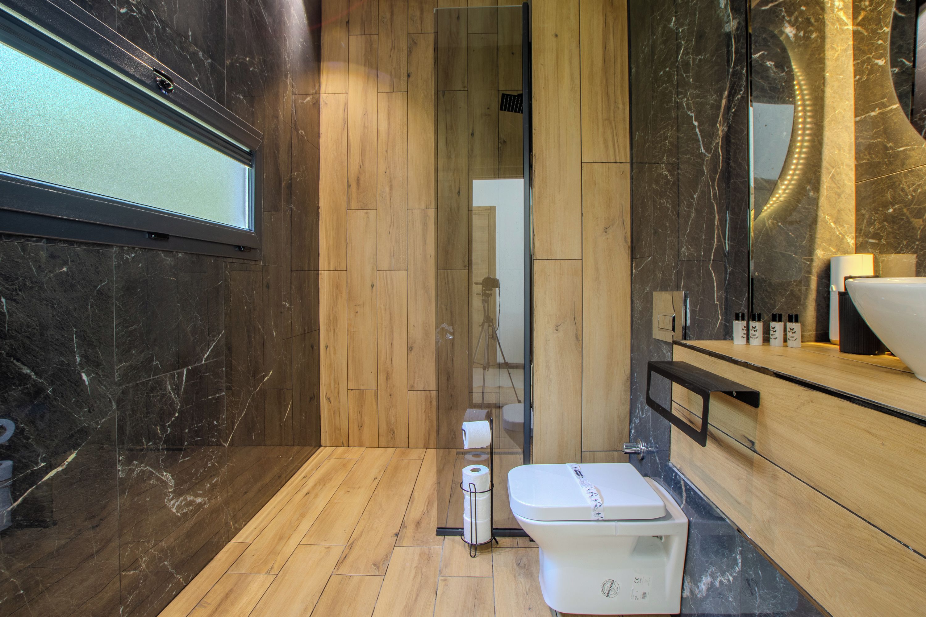 Een moderne badkamer met een houten vloer, een wit toilet, een douchecabine van glas en een marmeren wand.