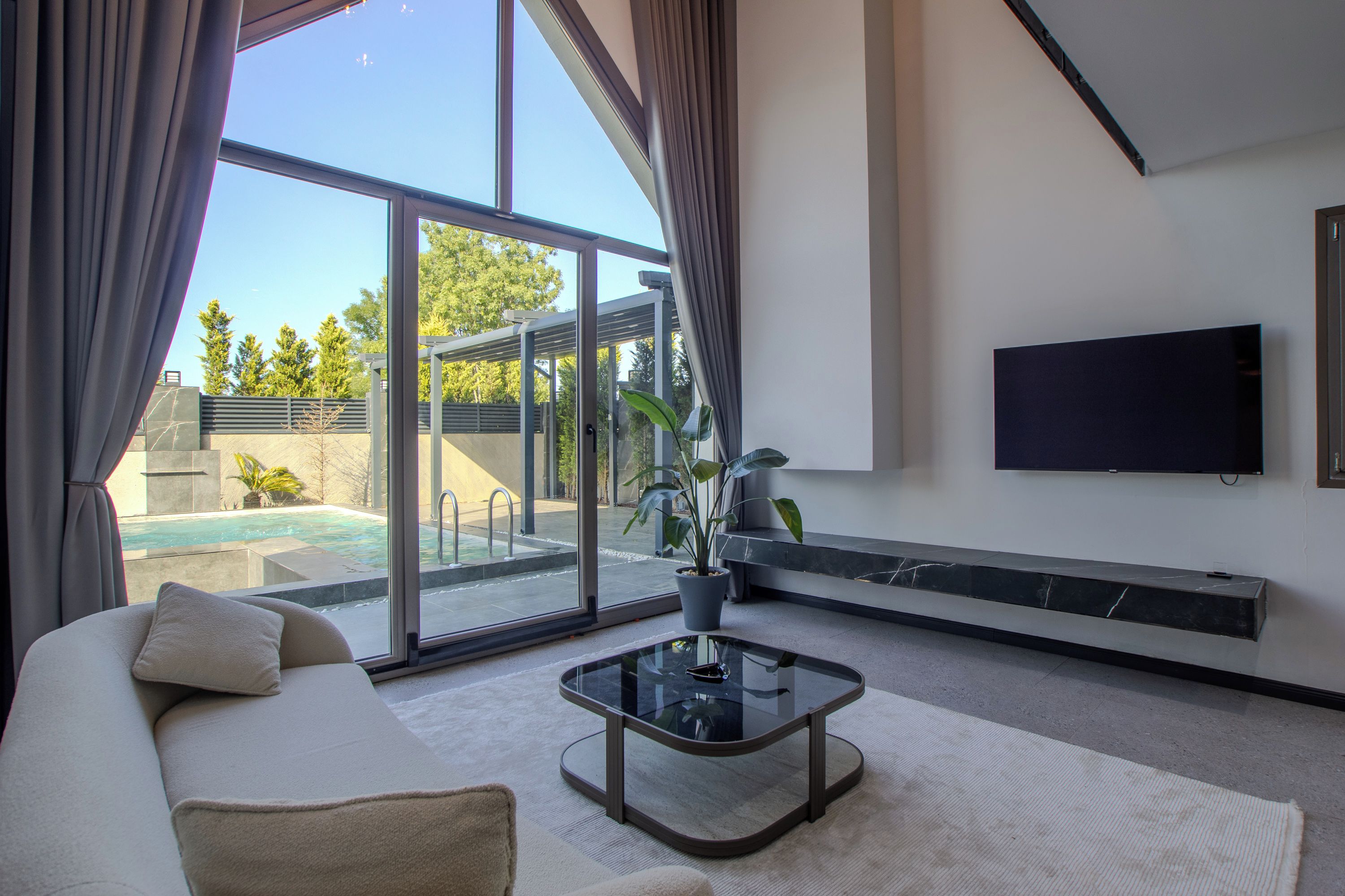 A modern living room with a sofa, a glass coffee table, a flat-screen TV mounted on the wall, and a view of an outdoor swimming pool.