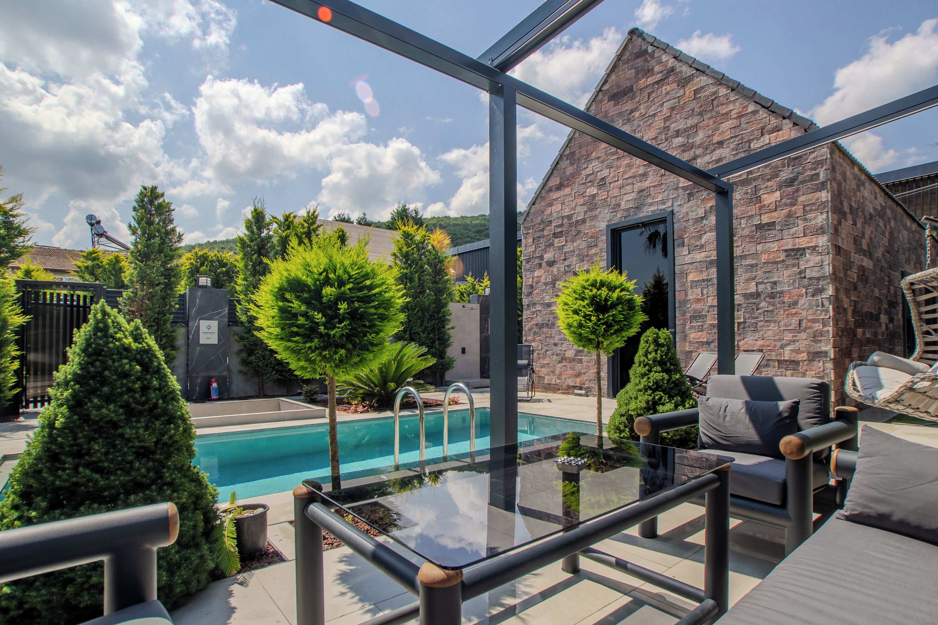 A modern outdoor pool area with a glass balustrade, stone building, and various greenery.