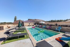 A swimming pool with lounge chairs and umbrellas, surrounded by a stone wall and a building with a tiled roof.