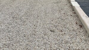 A building with a gravel driveway, a metal roof, and a paved area with a bench.