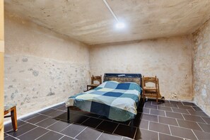A small, dimly lit room with a single bed, a chair, and a tiled floor.