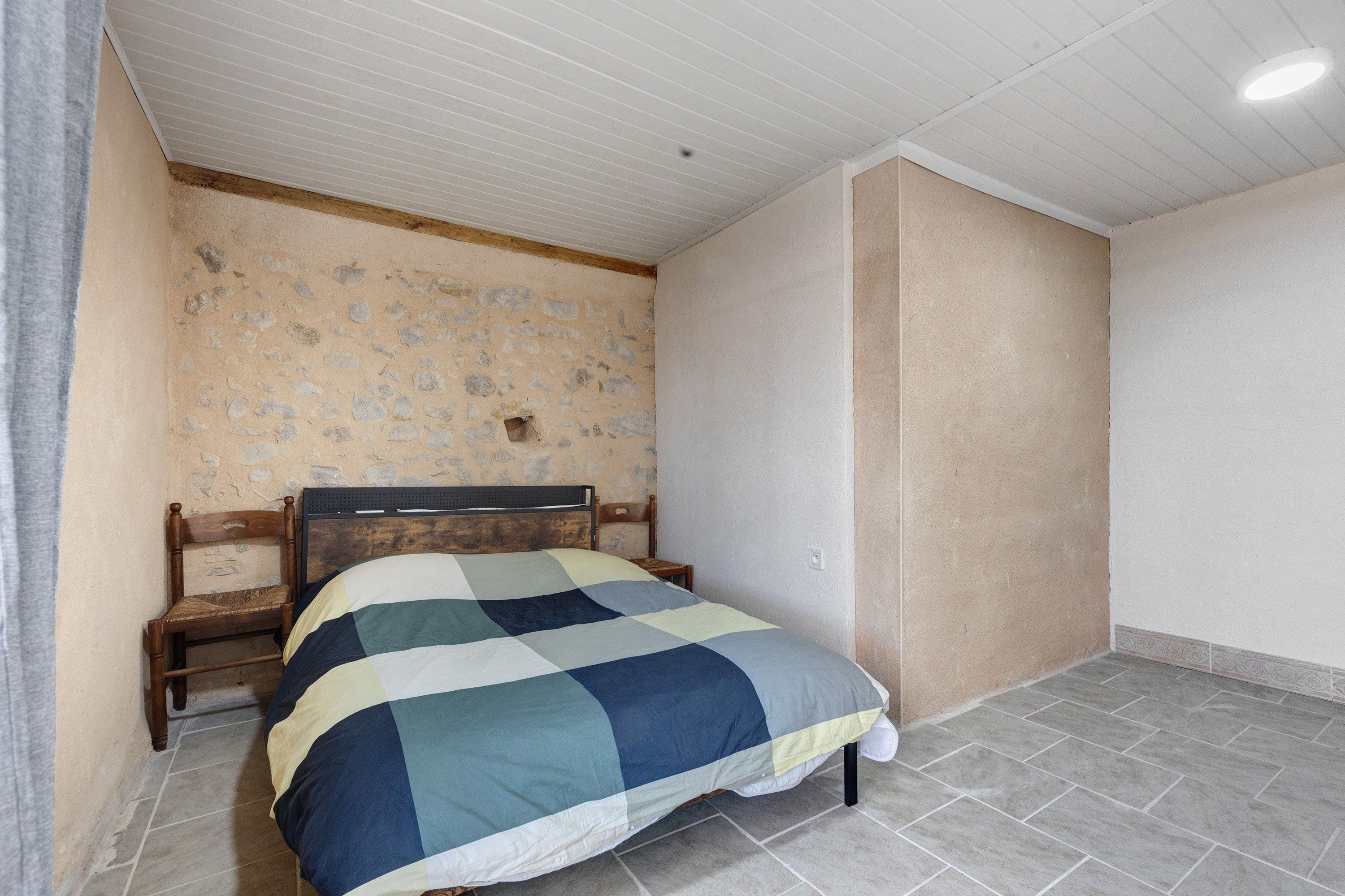 Quarto com uma cama, uma cadeira de madeira, parede de pedra e chão de azulejos.