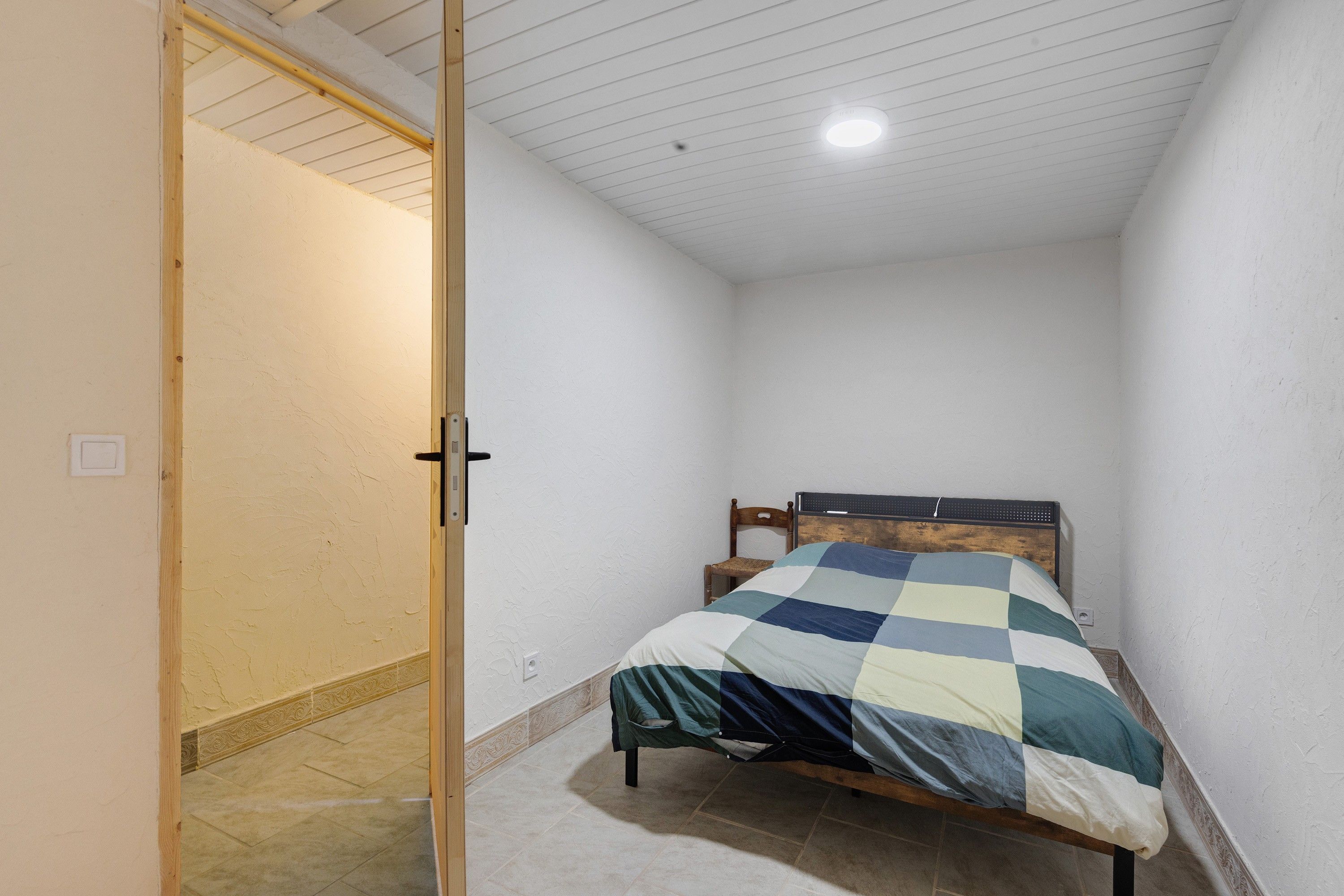 A small, narrow room with a bed, a door, and a ceiling light.