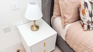 Una mesita de noche blanca con una lámpara, una cama con un cojín estampado y un cabecero gris.