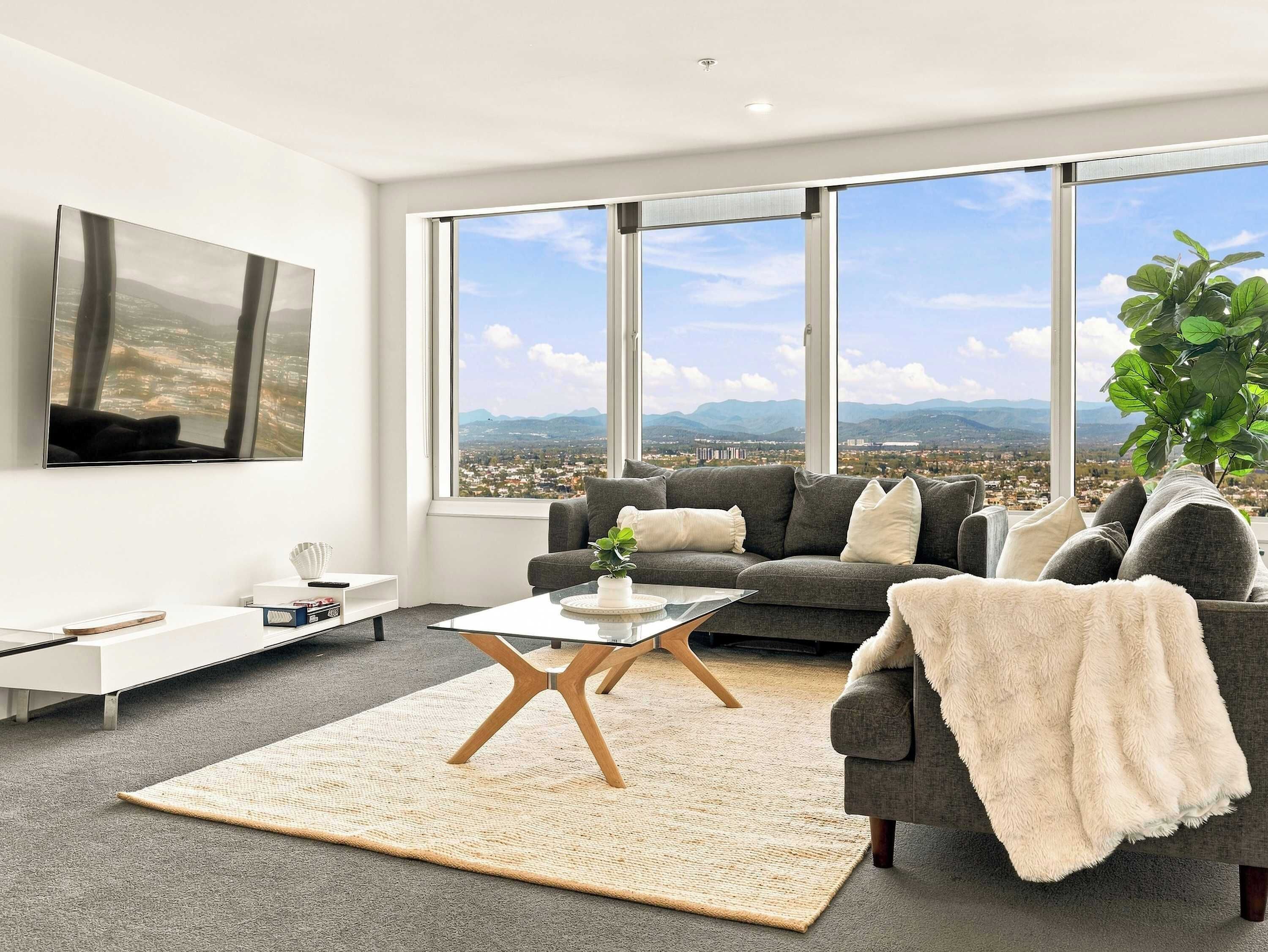 A modern living room with a large window, a grey sofa, a coffee table, and a TV mounted on the wall.