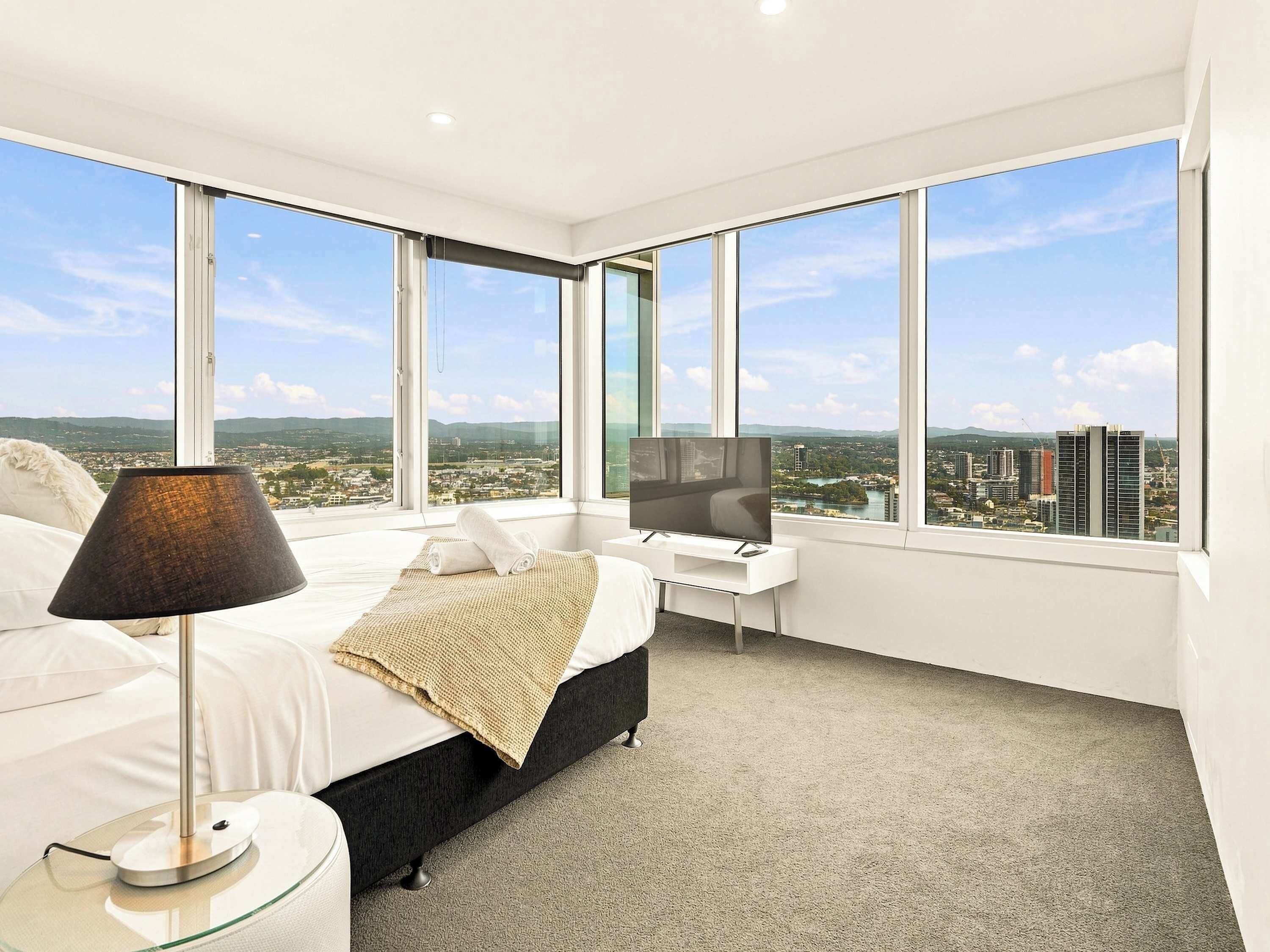 A modern hotel room with a large bed, a bedside table, a TV, and a view of a cityscape through large windows.