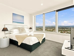 Ein Schlafzimmer mit einem Bett, zwei Nachttischlampen, einem Fernseher und einem großen Fenster mit Blick auf die Stadt.