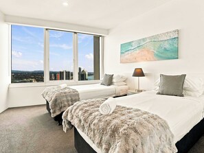 Quarto de hotel com duas camas, vista para a cidade, um quadro de uma praia e uma luminária.