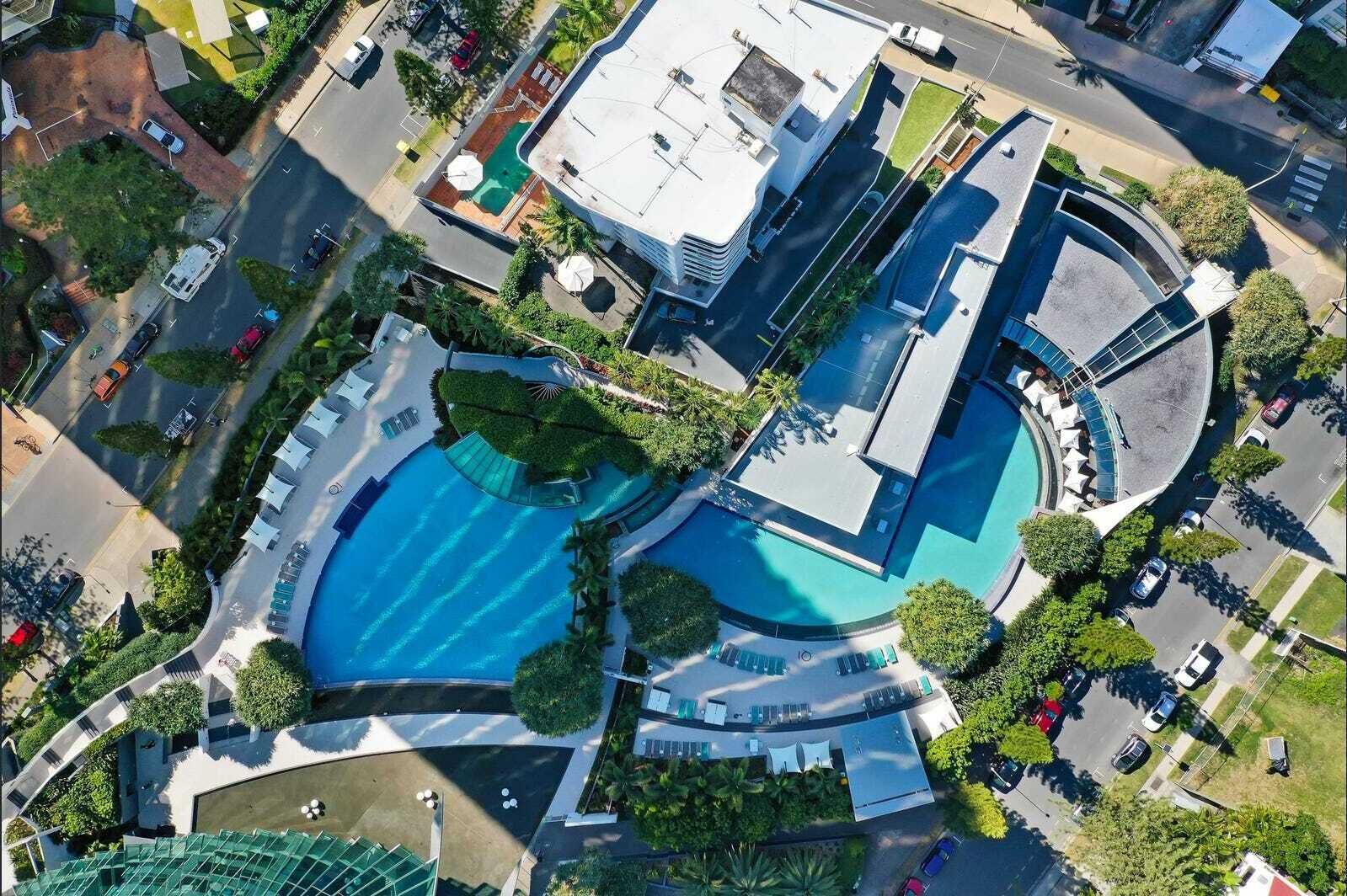 An aerial view of a modern hotel with a central swimming pool, multiple balconies, and a landscaped garden.