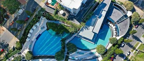 An aerial view of a modern hotel with a central swimming pool, multiple balconies, and a landscaped garden.