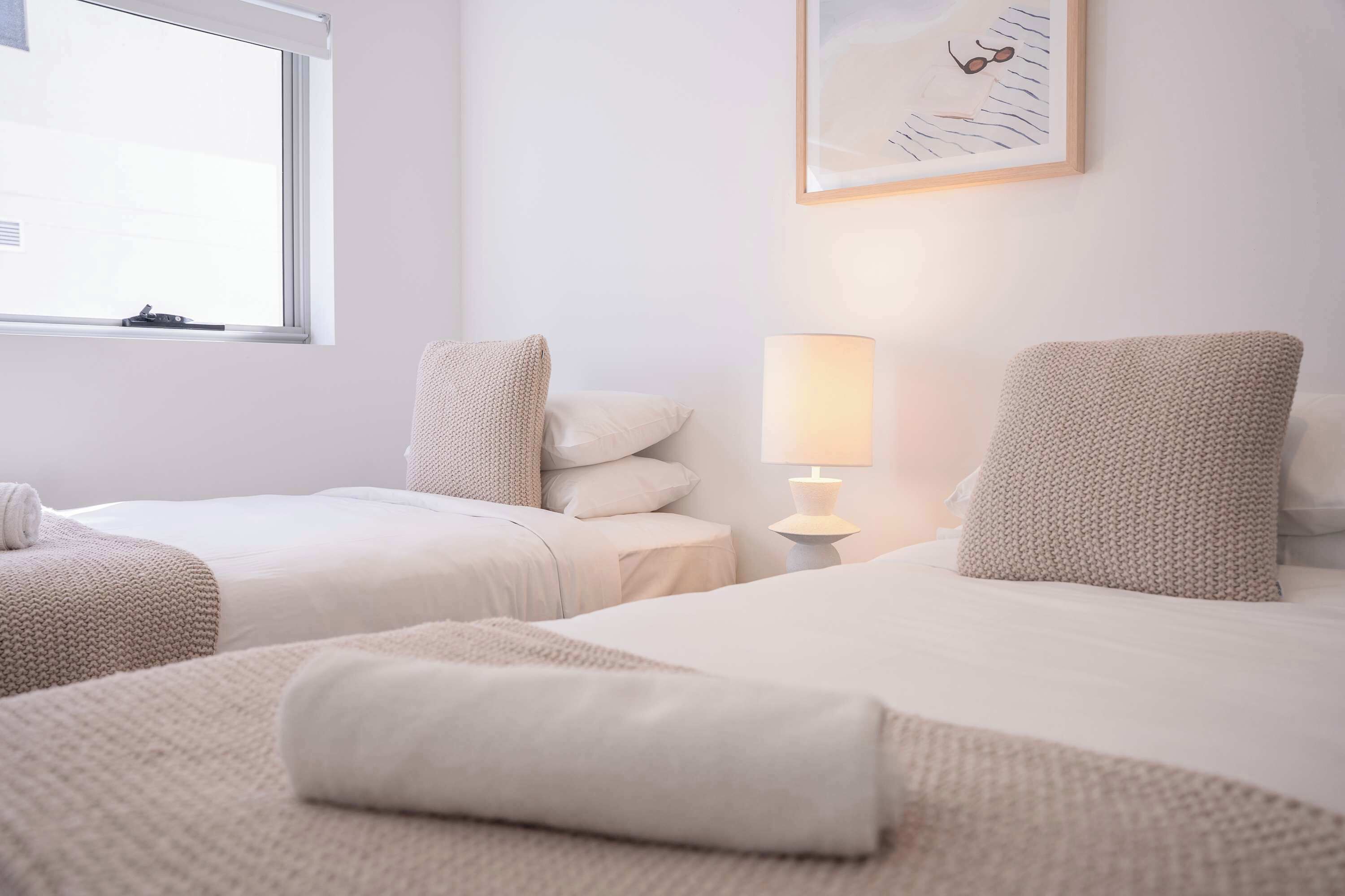 A hotel room with two beds, a window, a lamp, and a painting on the wall.