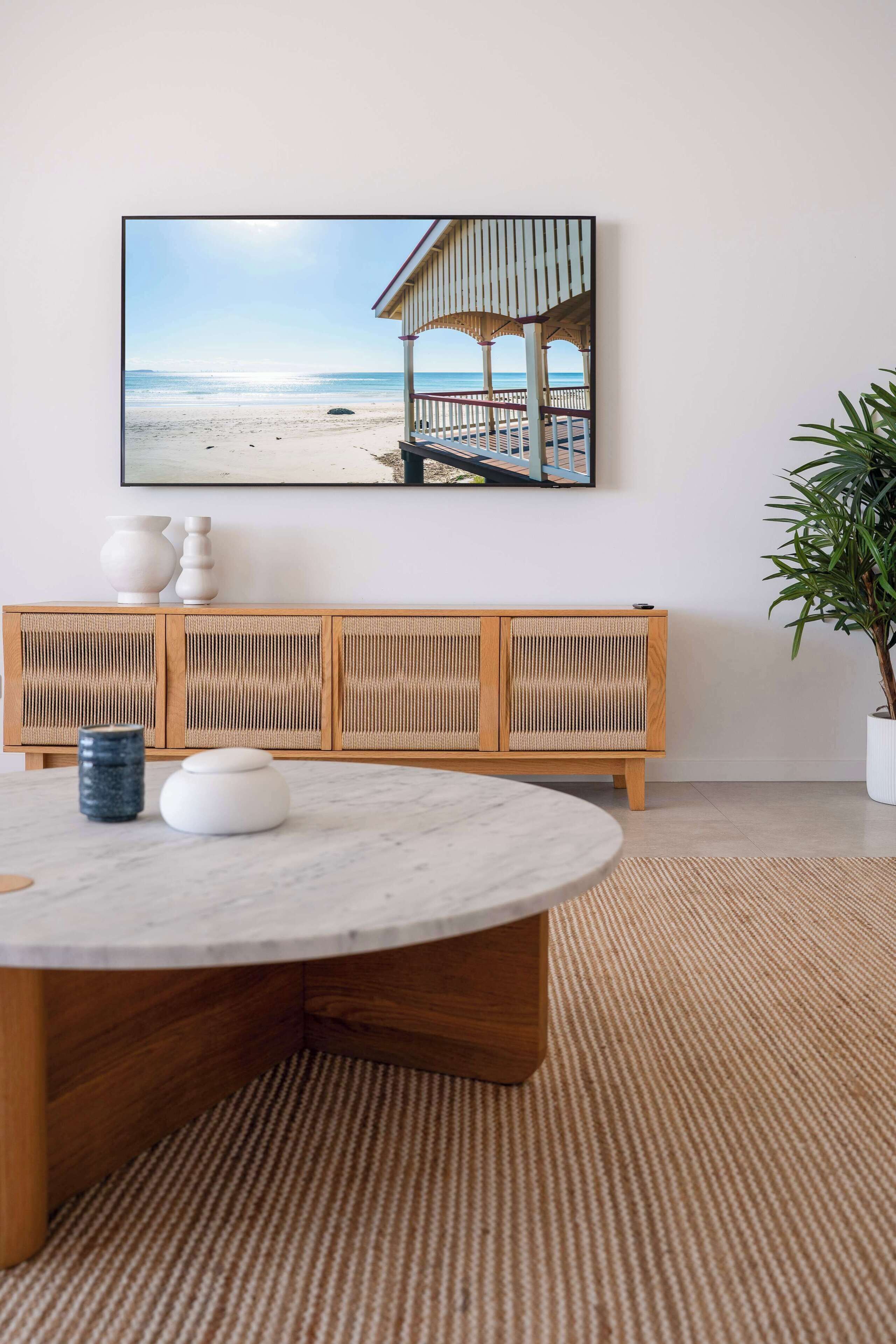 Ein Zimmer mit einem runden Tisch, einer Holzbank mit geflochtenem Rücken, einer Topfpflanze und einem Bilderrahmen mit Strandmotiv.