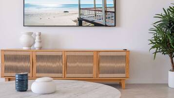 Une pièce comprenant une table ronde, un banc en bois avec un dossier en osier, une plante en pot et un tableau encadré représentant une scène de plage.