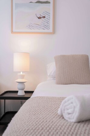Ein ordentlich bezogenes Bett mit einem weißen Kopfkissen und einer gemusterten Decke, eine Nachttischlampe und ein gerahmtes Bild an der Wand.