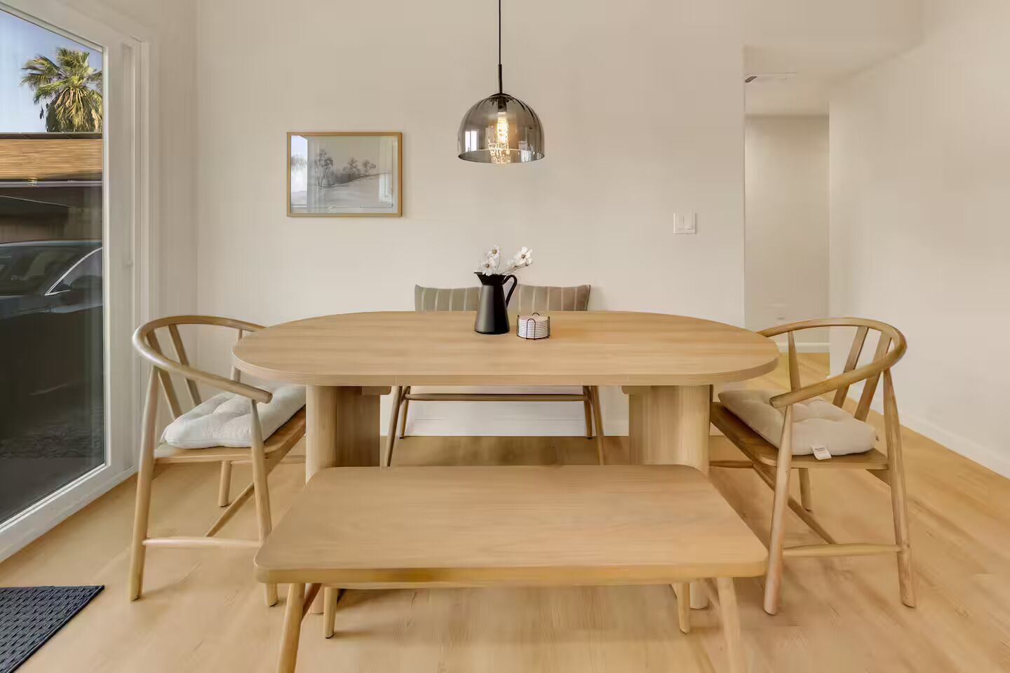 A dining area with a round wooden table, four chairs, a bench, a pendant light, and a framed picture on the wall.