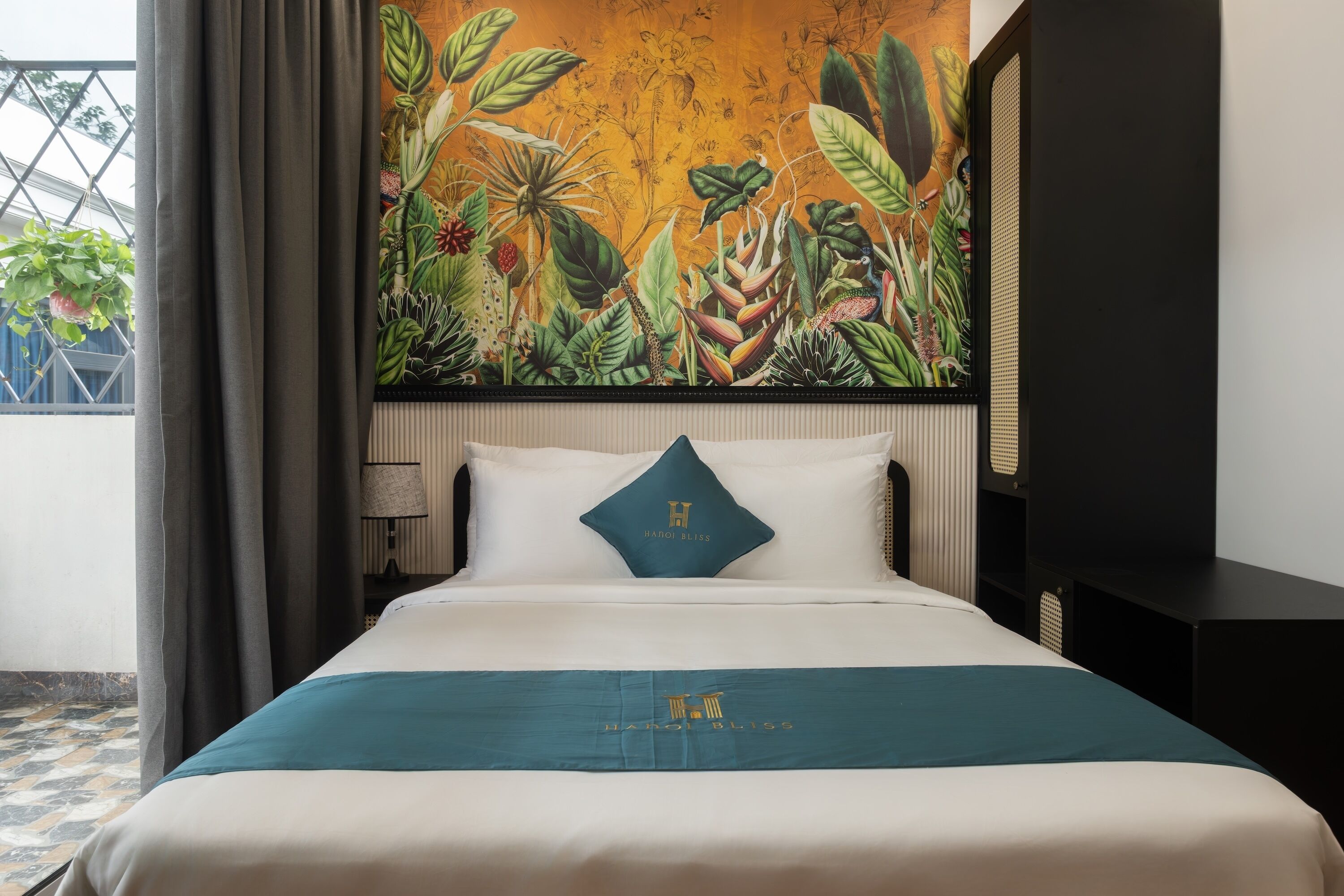 Ein Hotelzimmer mit Bett, Nachttisch, Lampe, einem Fenster mit Vorhängen und einem großen Wandbild mit tropischem Motiv.
